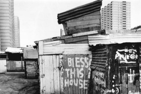 Black and white photos of shacks bunched together with buildings in the background