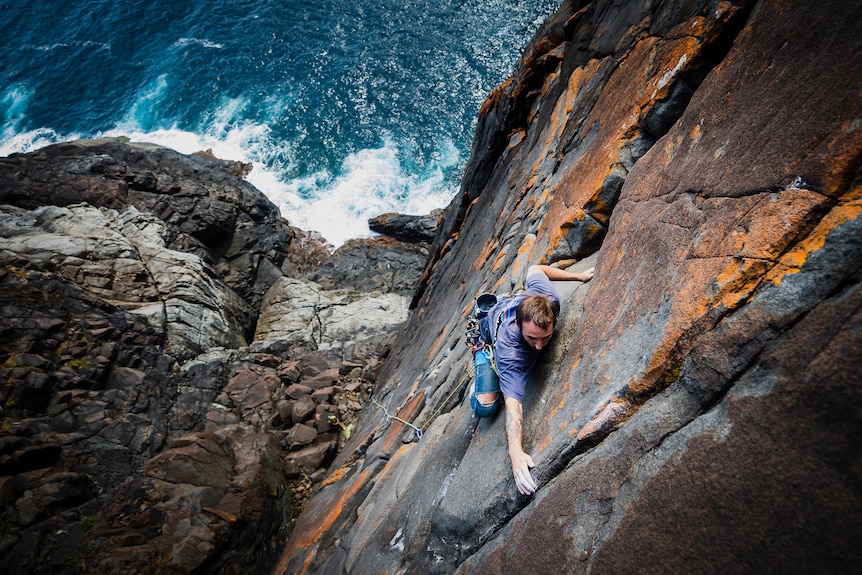 A man climbing up a cliff with the sea below.
