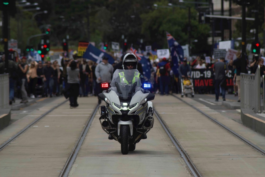 A police officer on a motorbike in focus with a line of protesters in the background.
