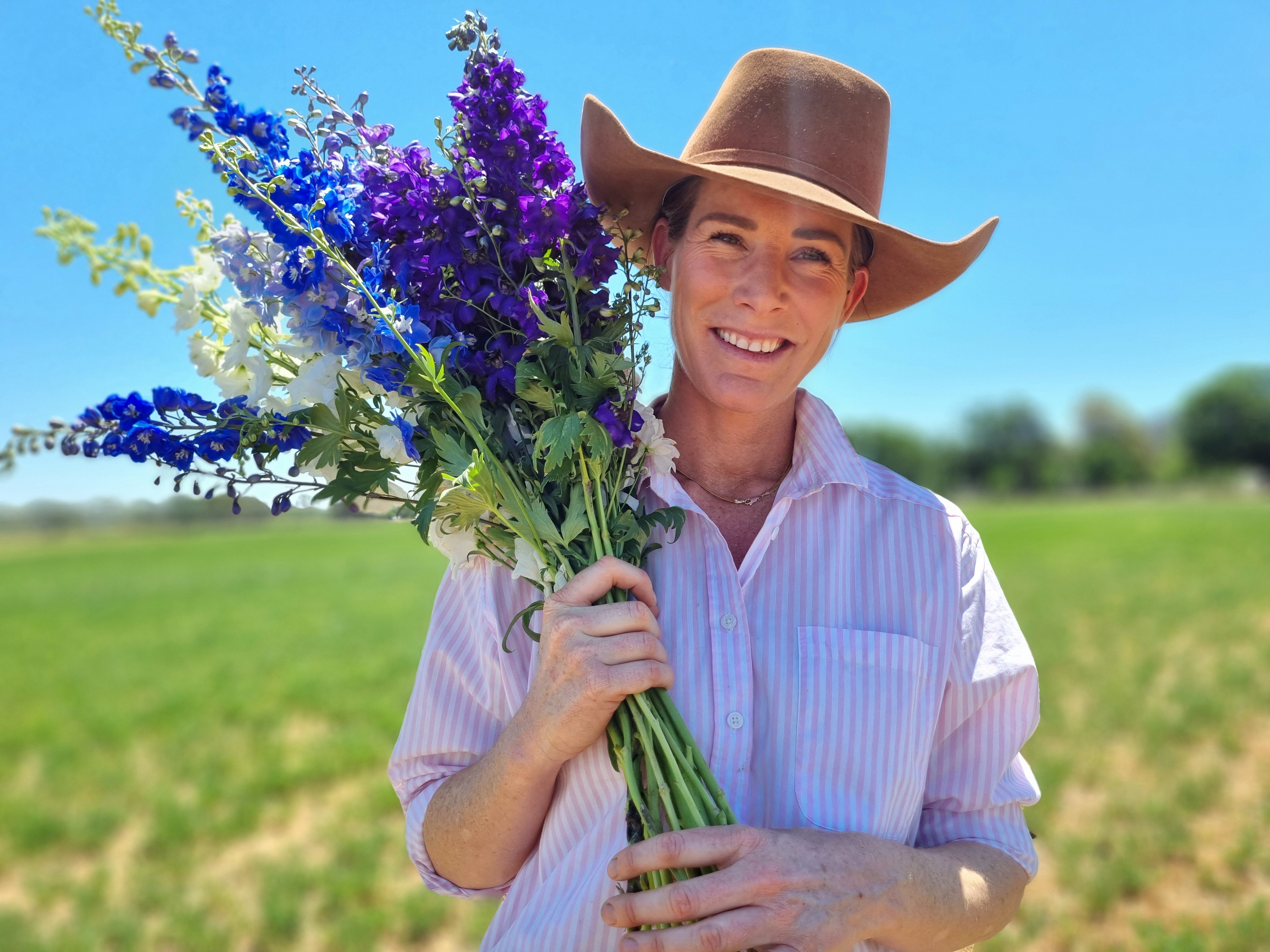 A woman in a hat stands in a paddock holding a bunch of purple and blue flowers.