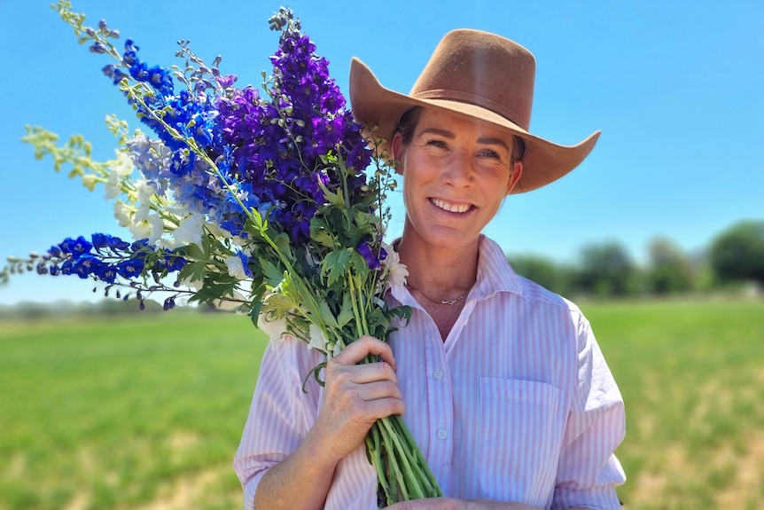 A woman in a hat stands in a paddock holding a bunch of purple and blue flowers.
