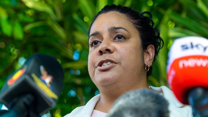 A woman with dark hair speaks into microphones. She is outside, and her expression is somber.