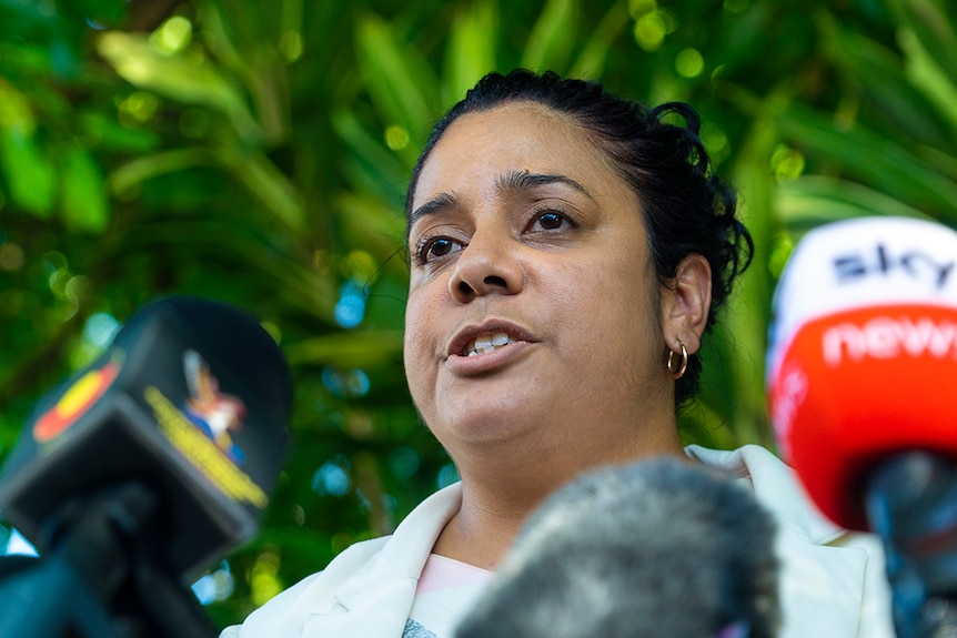 A woman with dark hair speaks into microphones. She is outside, and her expression is somber.