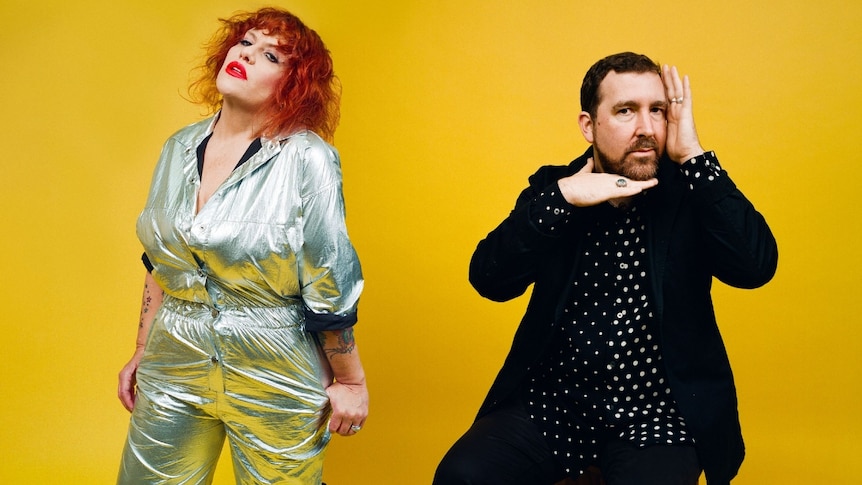 Amy Douglas wears a silver jumpsuit, Joe Goddard wears a polka dot shirt and black jacket. They stand before a yellow background