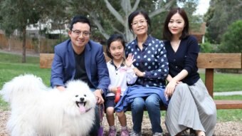 Sean Dong, his wife, mother and young daughter sit on a park bench smiling with their fluffy white dog