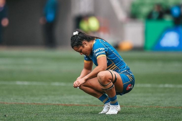Woman in blue uniform crouches down on rugby field. 