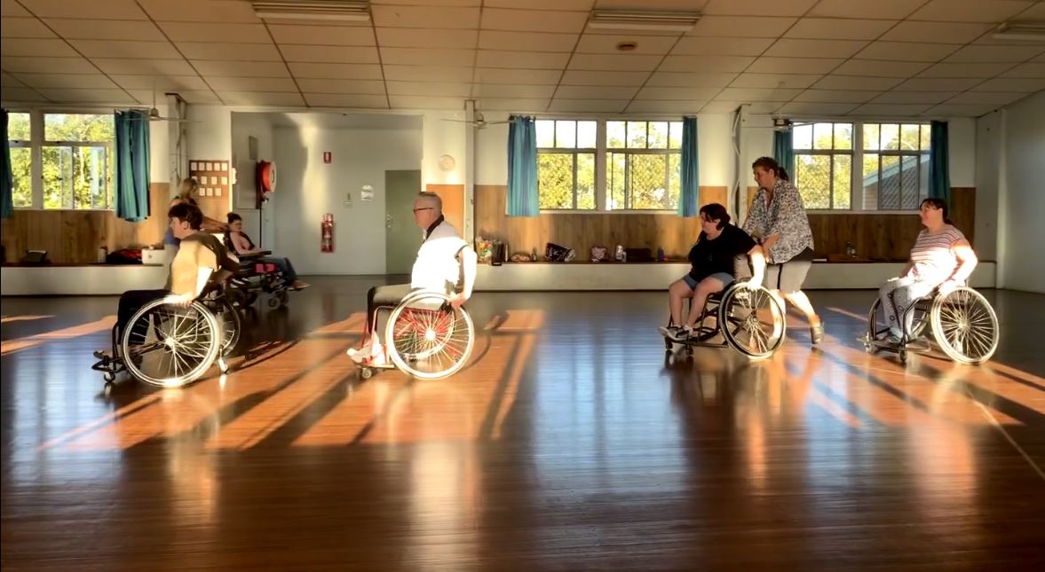 People in wheelchairs at a dancing rehearsal in a big room with a wooden floor.