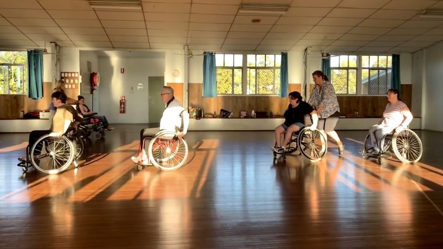 People in wheelchairs at a dancing rehearsal in a big room with a wooden floor.