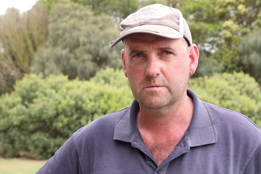 A man with a hat on stands looking into the camera with bushes in the background.