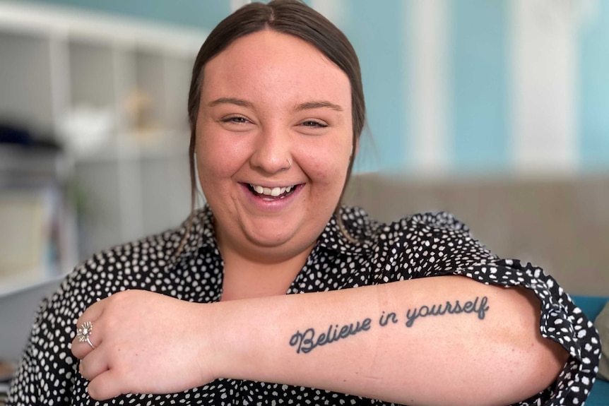 Young woman holds up forearm with 'Believe in yourself' tattooed on it.