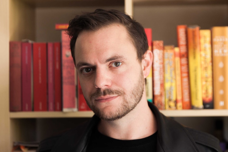 The writer Liam Pieper, leather jacket, beard, small smile, in front of a bookshelf