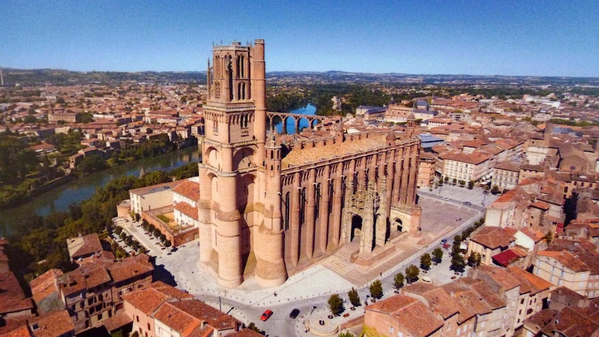 A colour film photo shows an aerial photo of the red-brick Albi Cathedral dominating a medieval town situated by a river.