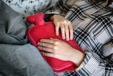 A person wearing a check shirt, lying down with a red hot water bottle on their stomach.