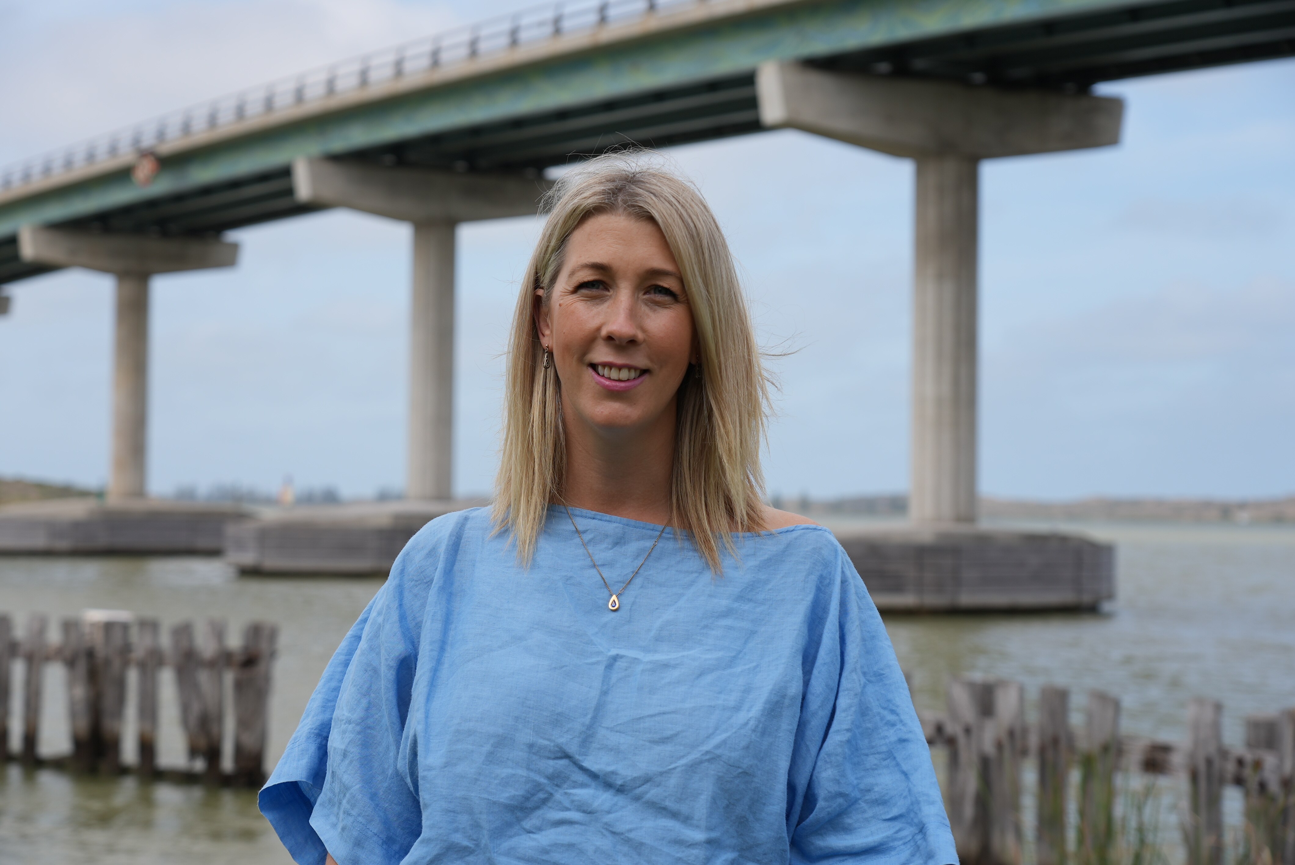 A blonde woman wearing a blue top in front of a bridge over a river