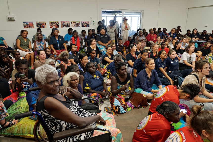 A group of mainly women sitting and standing inside a large room, ready to listen to someone speak.