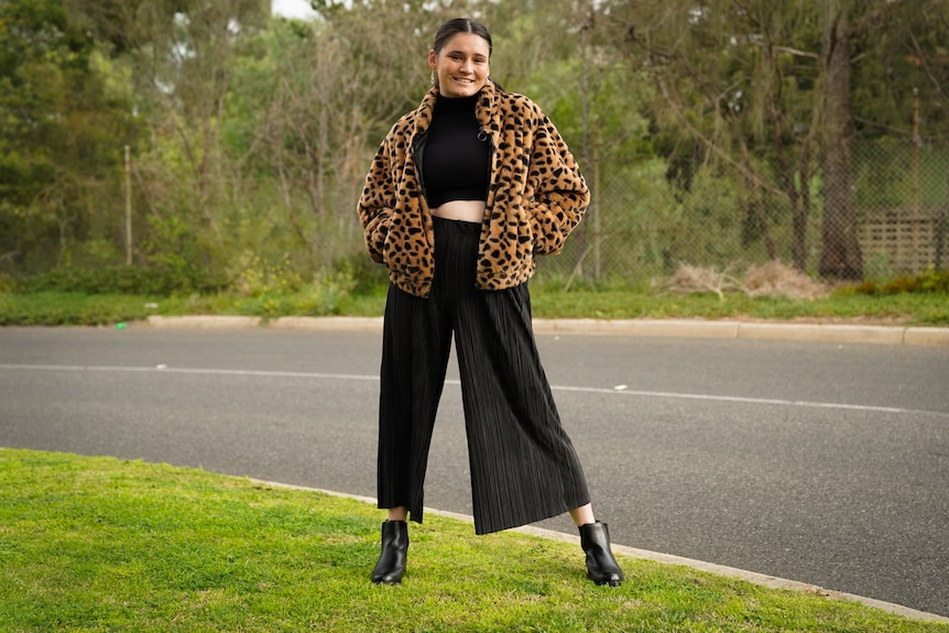 A far away picture sowing Molli Johns standing with one foot out, wearing black pants and top and leppard jacket. 