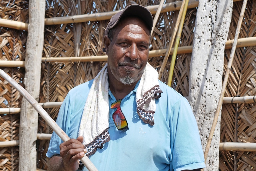 Man with hat and blue shirt, with towel around his neck holding a bamboo stick