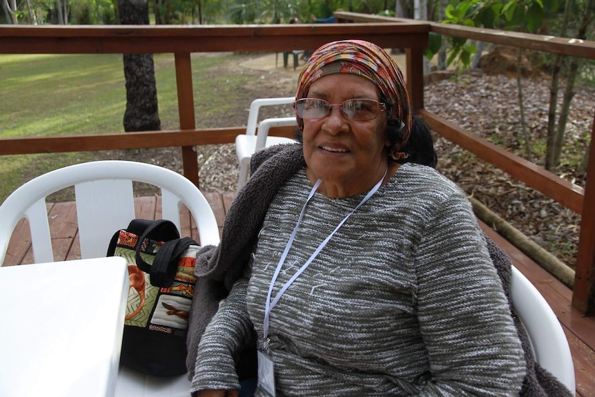 An older woman looks up at the camera from her chair in a garden.