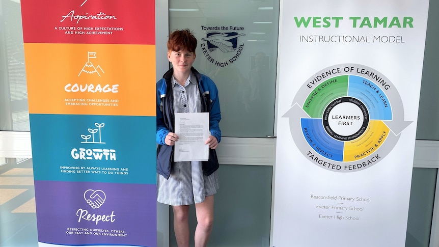 A young girl in a school uniform stands between two banners holding a letter.