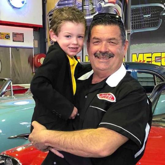 A man hold a young boy, both smiling, stand in front of two classic cars