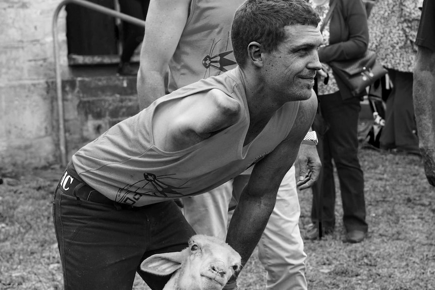 Josh Talbot holds a sheep down with his arm, looking ahead and smiling.