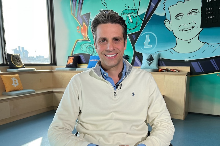 A Caucasian man with brown hair, sitting on a chair, smiling at the camera.