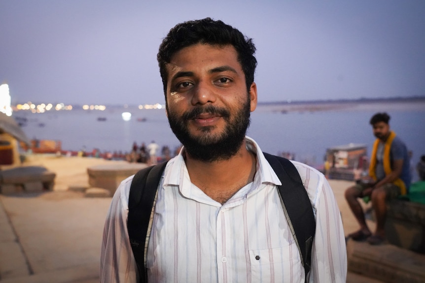 An Indian man wearing a white shirt and black backpack stands infront of a river.