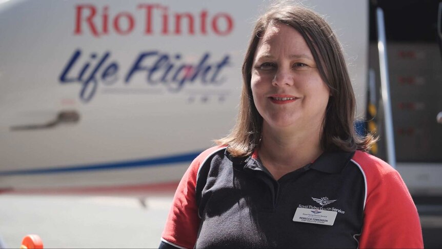 A woman with shoulder-length brown hair and a Royal Flying Doctor Service shirt smiles as she stands in front of an aircraft.