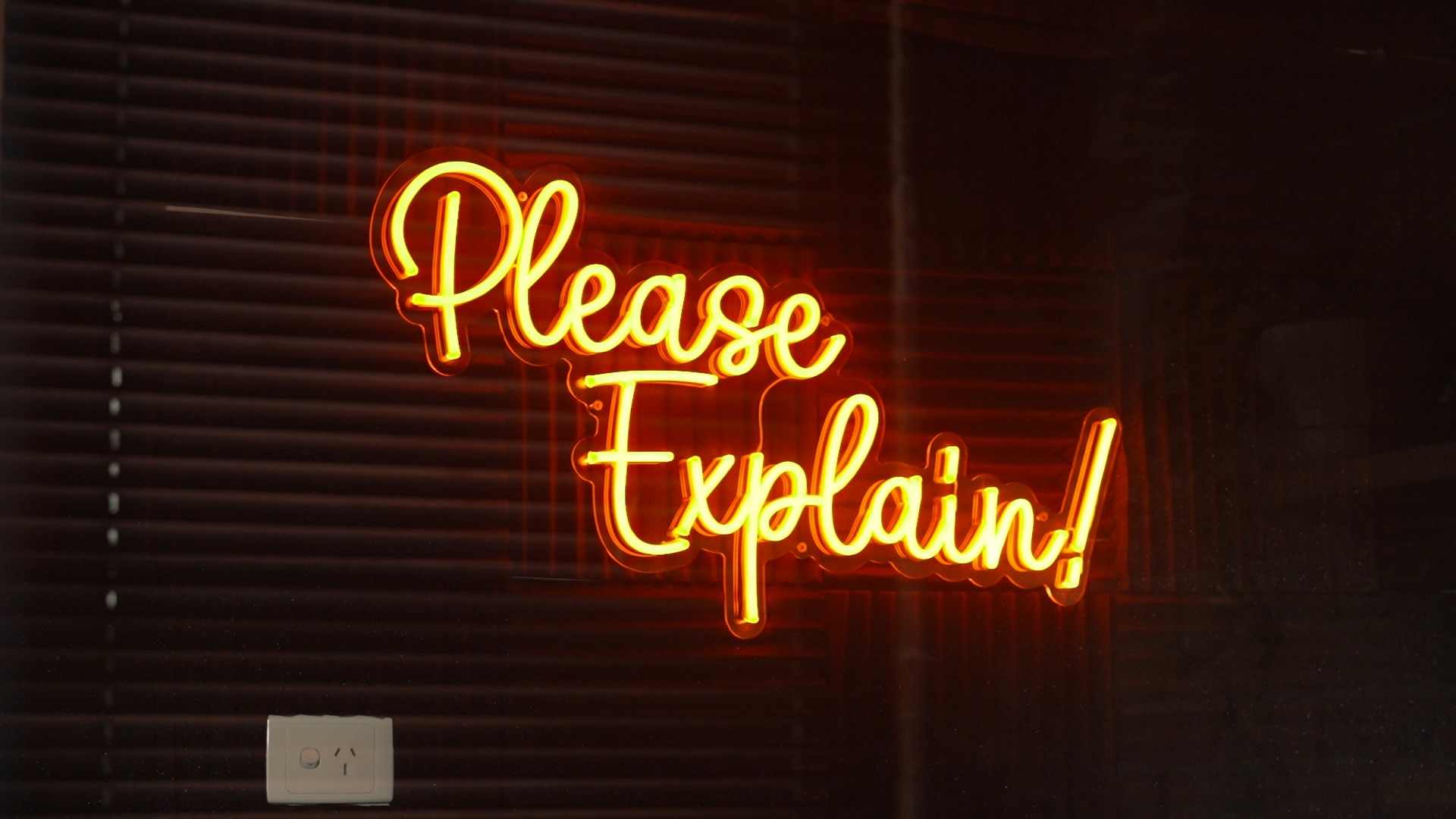 An orange neon sign that reads "Please Explain!" affixed to a wall in a dark room.