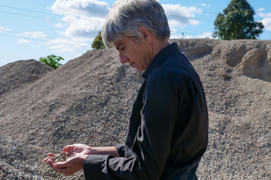 A woman with short, grey hair stands in a quarry and examines some rocks in her hands.