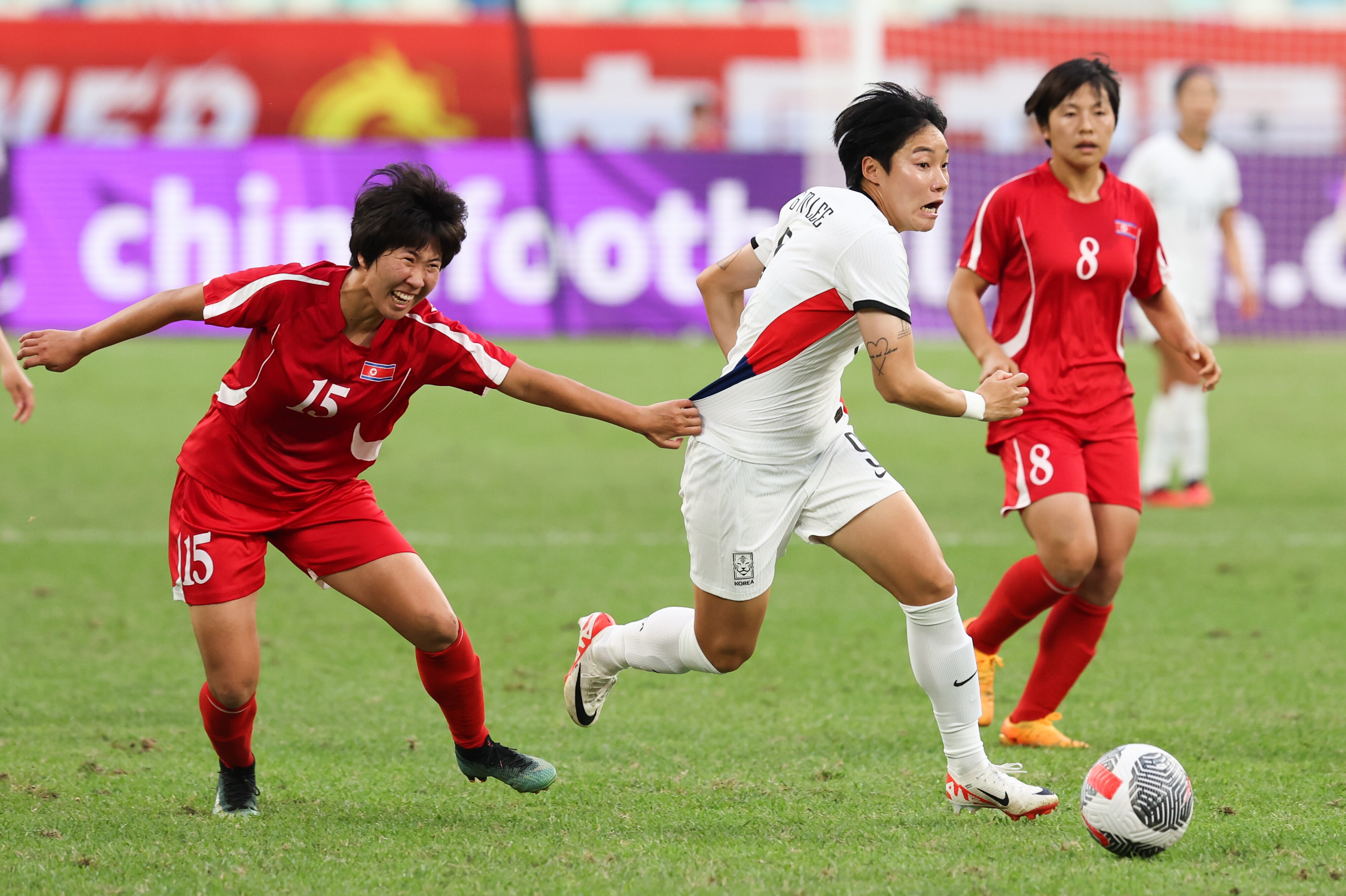 A soccer player wearing white runs with the ball while being held back by an opponent wearing red during a game