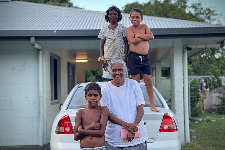 An older woman and three children stand around a car in front of a famiy home.