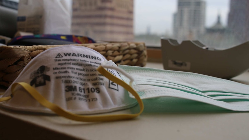 A P2/N95 mask and a surgical mask are placed on a desk in front of a window with a city skyline view.
