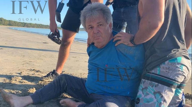 Peter Foster sits on the sand in handcuffs flanked by plainclothes police.