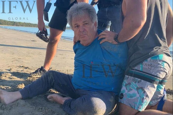 Peter Foster sits on the sand in handcuffs flanked by plain clothes police.