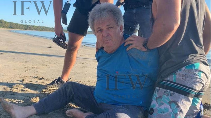 Peter Foster sits on the sand in handcuffs flanked by plain clothes police.