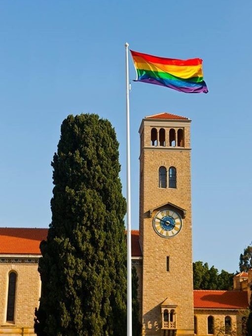 A rainbow flag flies in front of a building at the University of Western Australia against a blue sky.