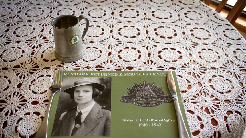 A placemat memorial to Elaine Balfour-Ogilvy on a table set with a doily tablecloth, cutlery and pewter mug.