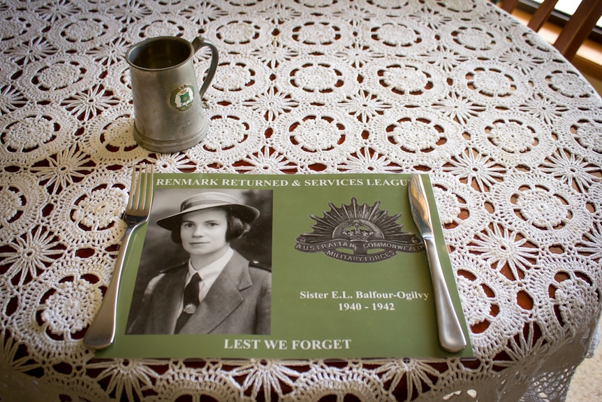 A placemat memorial to Elaine Balfour-Ogilvy on a table set with a doily tablecloth, cutlery and pewter mug.