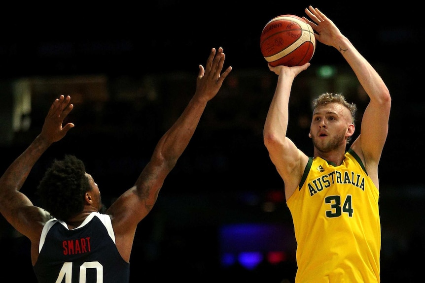 A male basketballer shoots for a basket with his right hand as an opponent attempts to block.