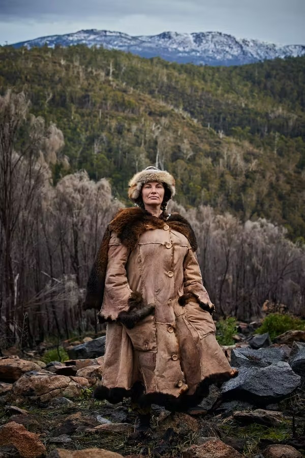a woman in a brown possum skin coat stands with grey sky, snowy mountains and bushland behind her