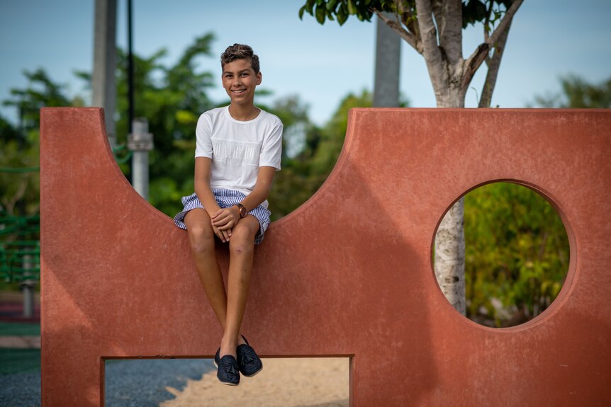 A young boy wearing a white tee shirt and blue shorts sits on a playground and is smiling.