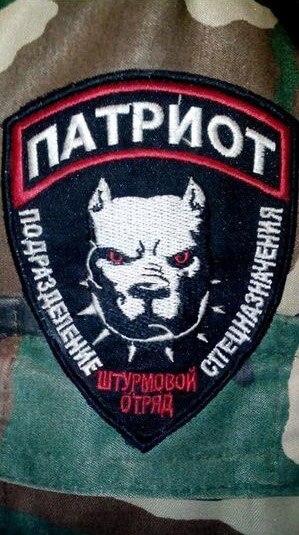 A close-up on a military patch sewn onto camouflage print shows a dog wearing a spiked collar, surrounded by Cyrillic letters