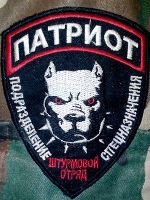 A close-up on a military patch sewn onto camouflage print shows a dog wearing a spiked collar, surrounded by Cyrillic letters