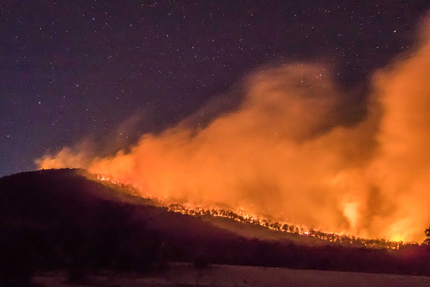 Flames burn over the ridge of a hill at night.