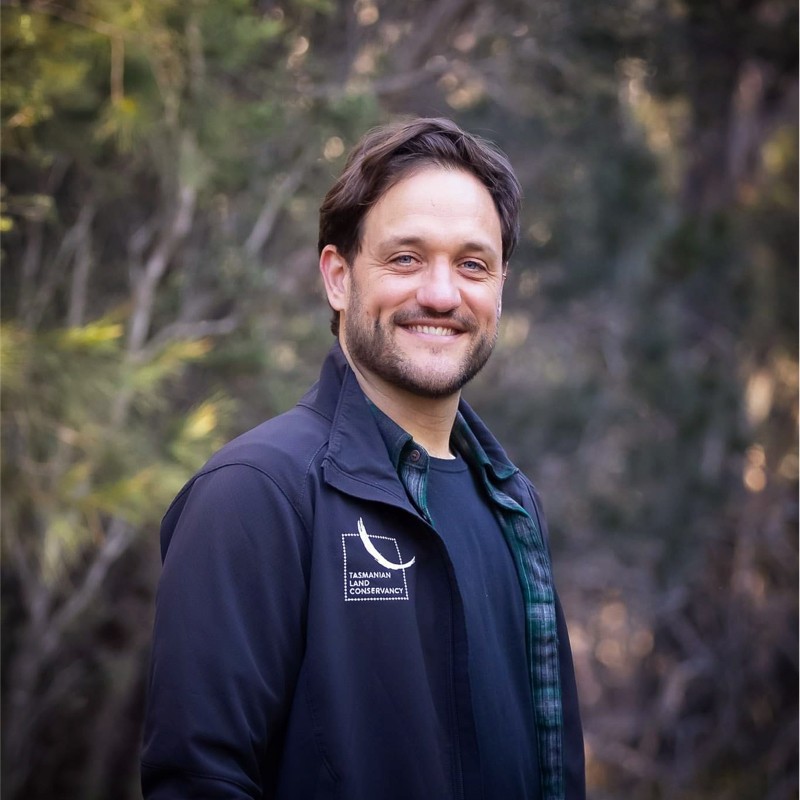 A man with dark hair and goatee smiles at the camera with bushland in the background.