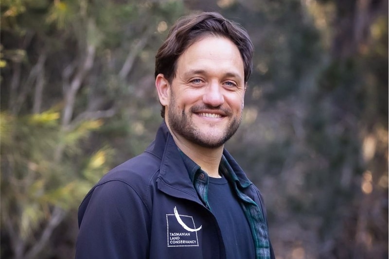 A man with dark hair and goatee smiles at the camera with bushland in the background.