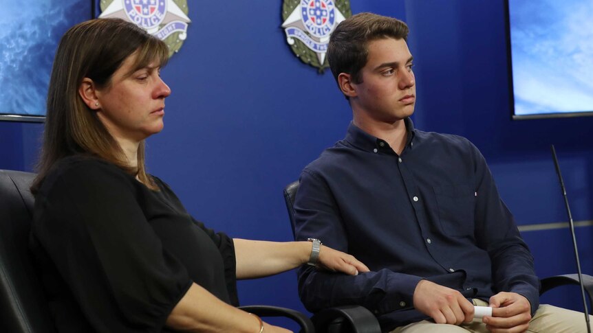 A woman who has been crying holds the arm of a young man as they sit in front of Victoria Police logos.