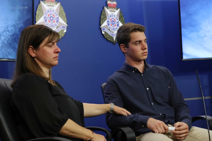 A woman who has been crying holds the arm of a young man as they sit in front of Victoria Police logos.
