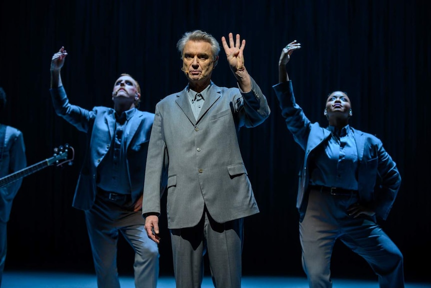 David Byrne in grey suit stands spot-lit on stage holding 4 fingers up, with two barefoot dancers in grey suits in background.